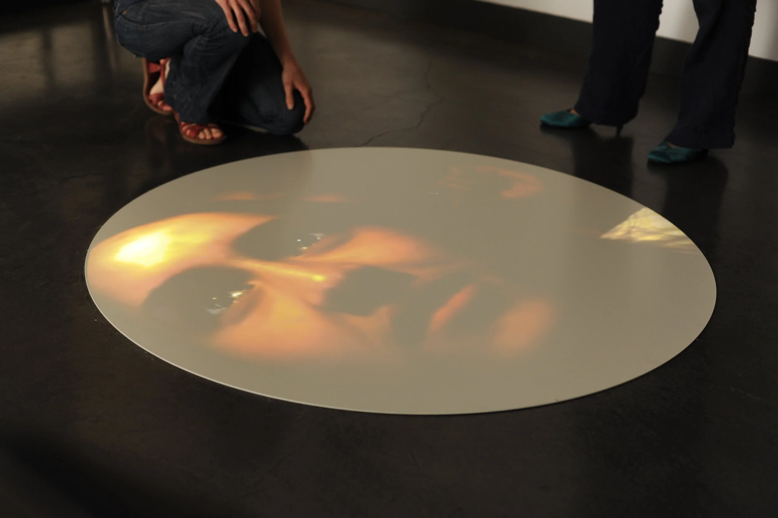 Une installation d'art numérique représentant une image lumineuse et orange sur une surface blanche, située au sol dans une galerie d'art, avec deux personnes observant l'œuvre, l'une en position accroupie et l'autre debout.
