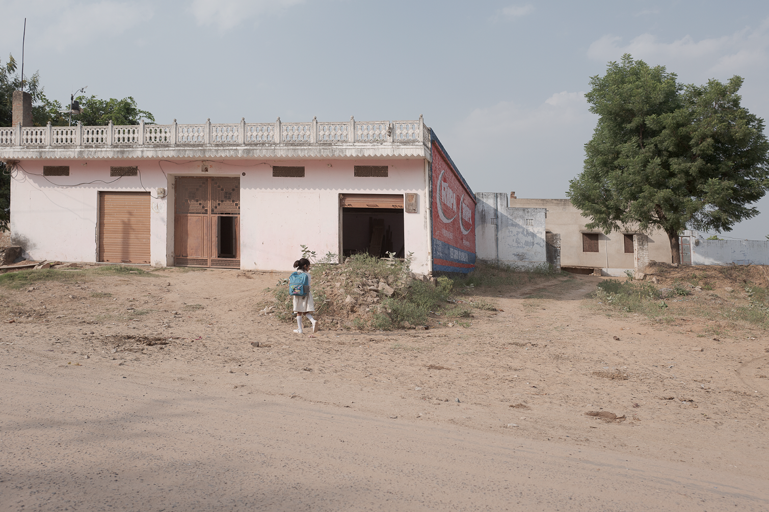 Une jeune fille avec un sac à dos blanc et bleu marche vers une maison simple en terre poussiéreuse, avec un arbre à droite et un mur avec une publicité Coca-Cola derrière.