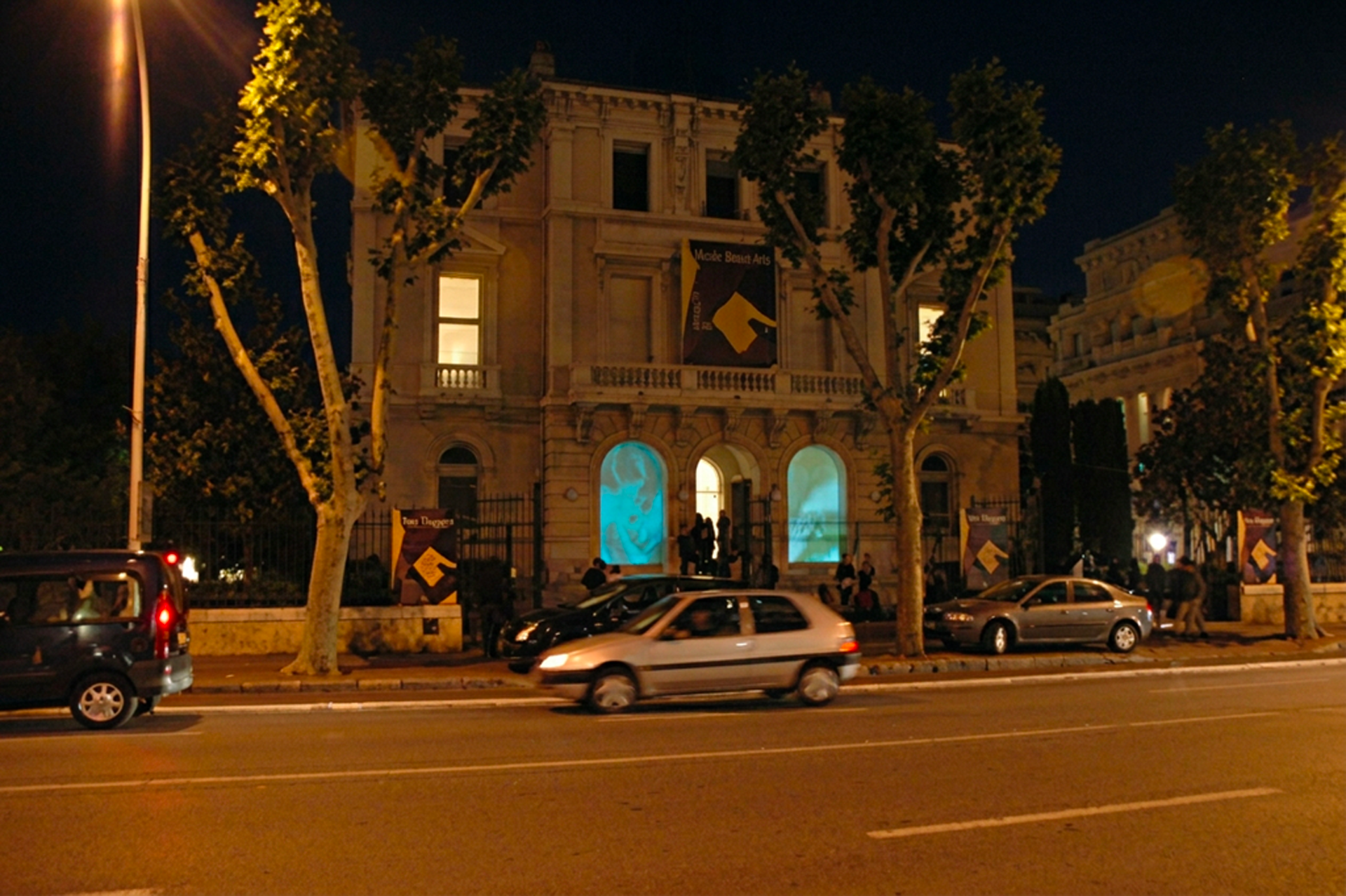 Vue nocturne d'un bâtiment historique éclairé avec des arbres en avant, des voitures sur la route et une projection bleue sur l'entrée.