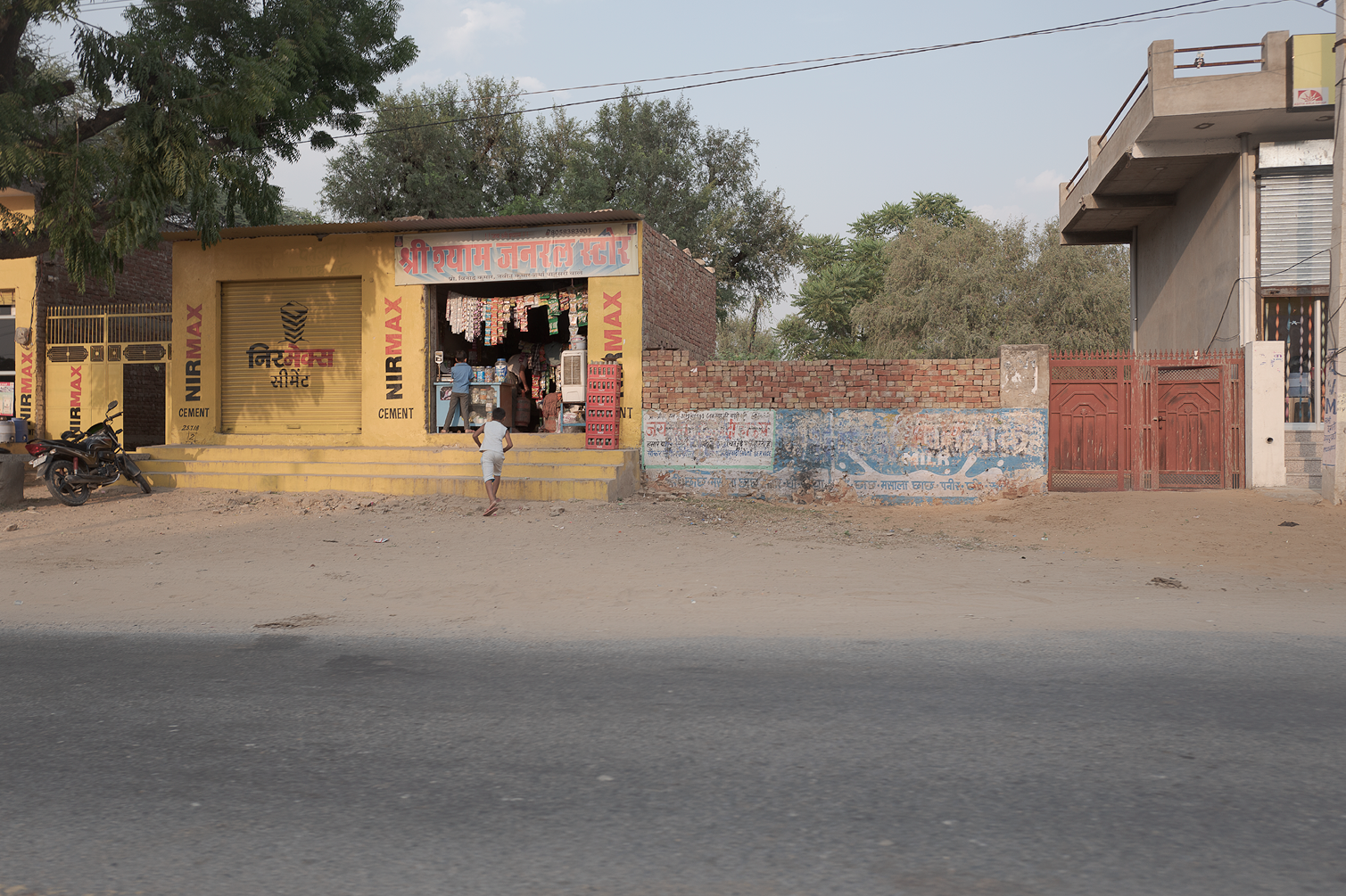 Un petit commerce en Inde avec une façade jaune, une escalerie menant au magasin, un mât à drapeaux, quelques enfants jouant dehors, un mur derrière avec des graffitis, une porte en métal rouge, et un environnement urbain.»