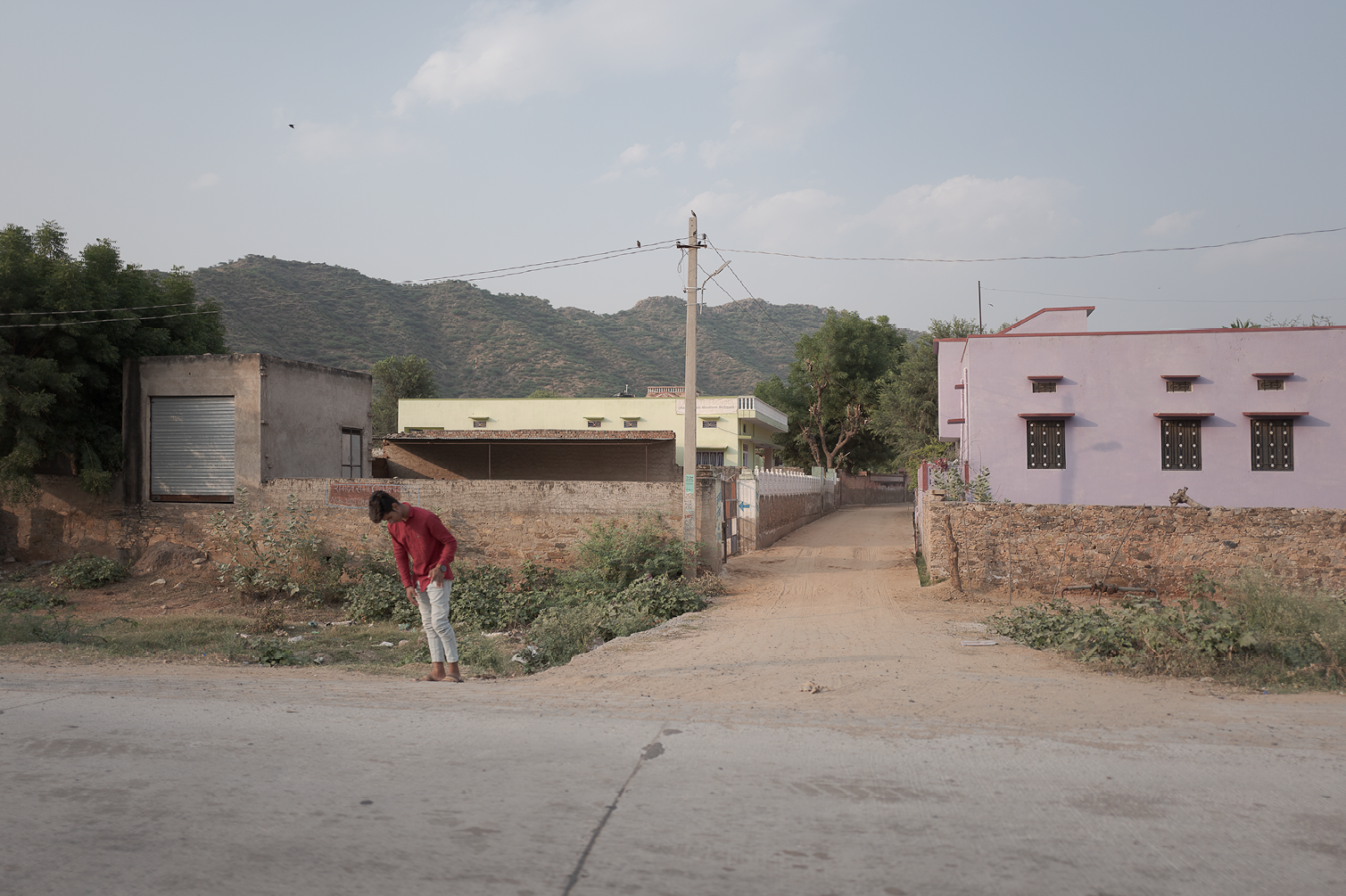 Une jeune personne en vêtements rouges et blancs marche le long d'une route en terre dans un village avec des maisons colorées et des montagnes en arrière-plan.