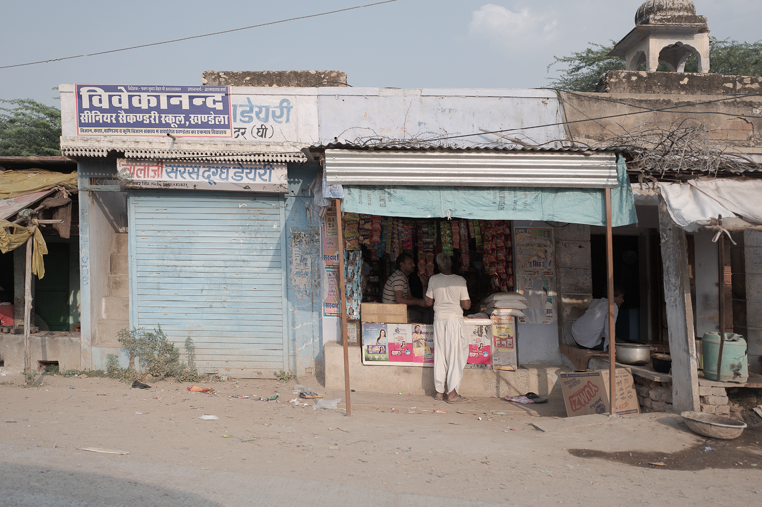 Une petite boutique en bord de route avec deux hommes à l'entrée. La boutique vend des snacks, des produits de base et des articles divers. La devanture est recouverte partiellement d'une toile bleue et le bâtiment semble usé, avec un bâtiment en pie