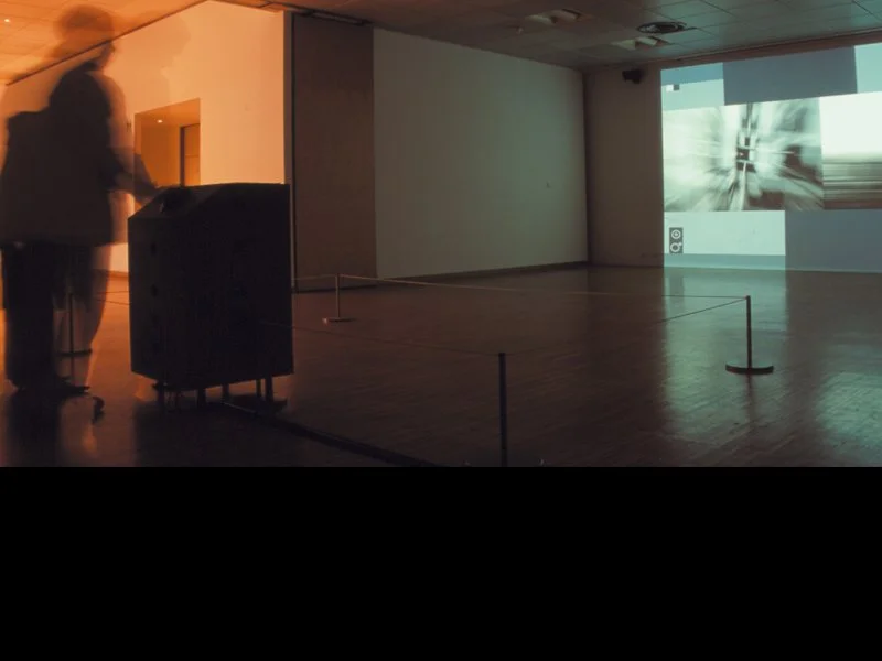 Une salle d'exposition ou de projection, avec une vidéo ou un film en cours de projection sur un mur blanc et une personne floue observant la scène.