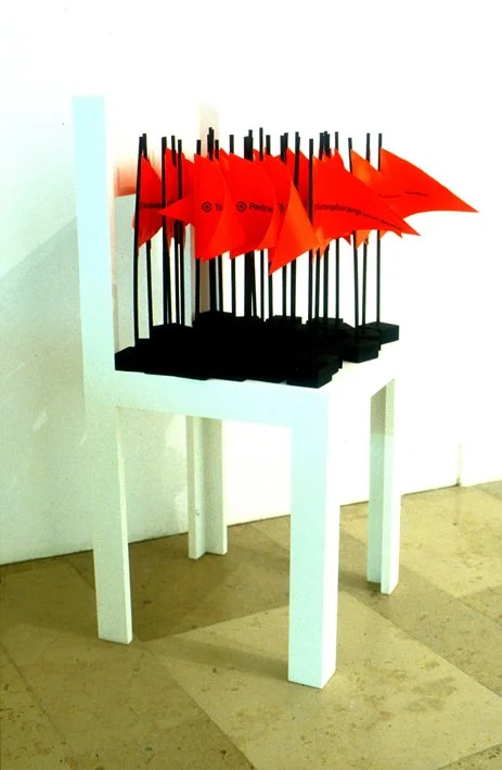 Installation artistique avec plusieurs petits drapeaux rouges sur des tiges noires, placée sur une table blanche. Bâtons noirs avec des drapeaux en tissu rouge, créant un contraste visuel.