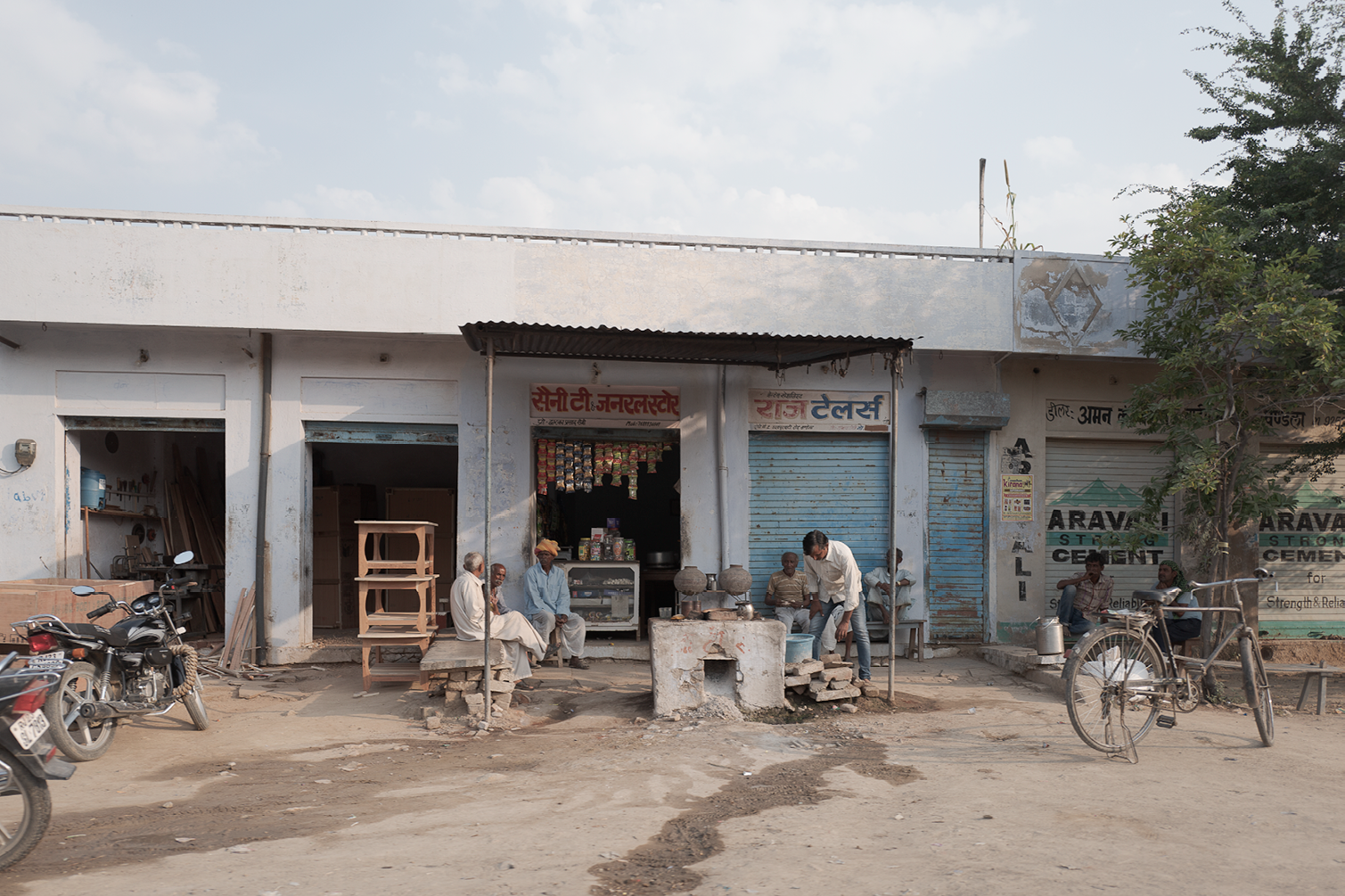 Vue d'une petite rue en Inde avec plusieurs hommes assis devant des boutiques, un vélo et des motos stationnés, et un arbre à droite, sous un ciel en partie nuageux.