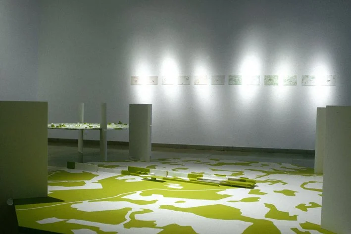 Exposition d'art contemporain dans une galerie, avec des œuvres sur le mur et une installation au sol composée de formes blanches et vertes.