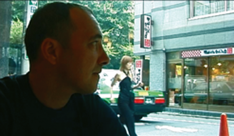 Un homme assis dans un café en ville avec des immeubles et des passants en arrière-plan.