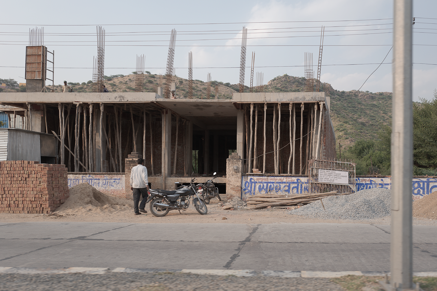 Construction d'un bâtiment avec des gens dessus, scooter garé devant, piles de briques et matériaux de construction devant un mur.
