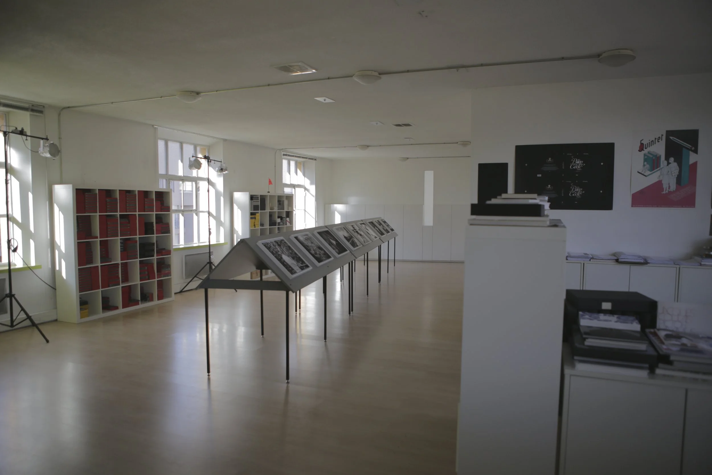 Salle d'exposition avec des présentoirs contenant des photographies et des posters, luminaires suspendus, fenêtres laissant entrer la lumière naturelle, étagères avec des livres ou des catalogues, ambiance claire et organisée.