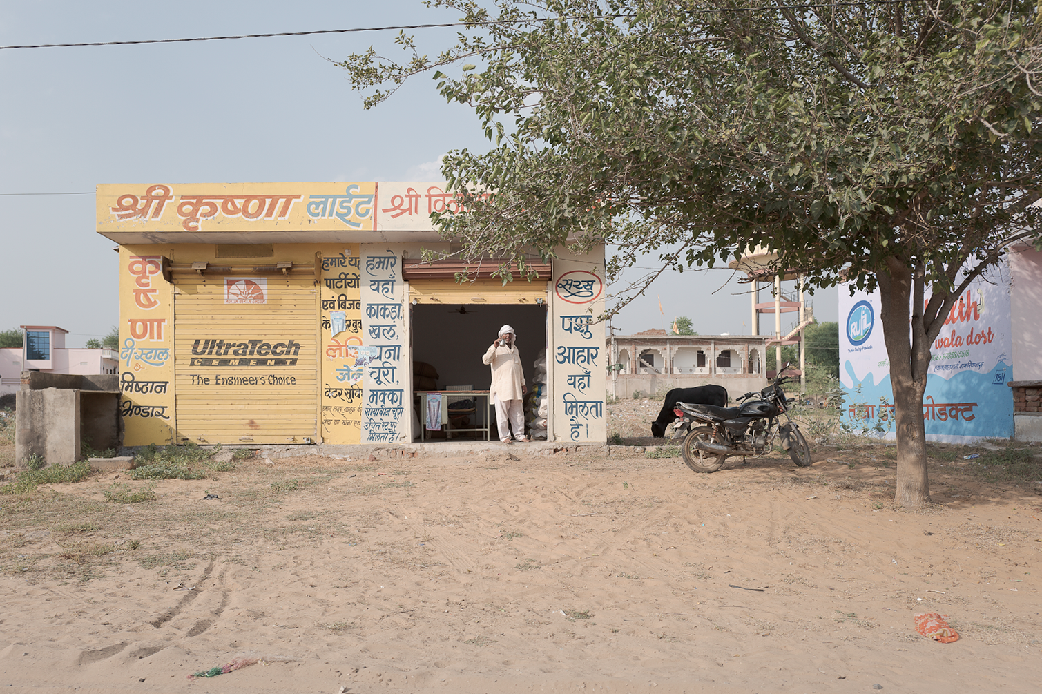 Une petite boutique avec des panneaux en hindi et des inscriptions en anglais, une personne parlant au téléphone, un scooter garé à côté d'un arbre en dehors d'une zone urbaine, avec un bâtiment en construction en arrière-plan.