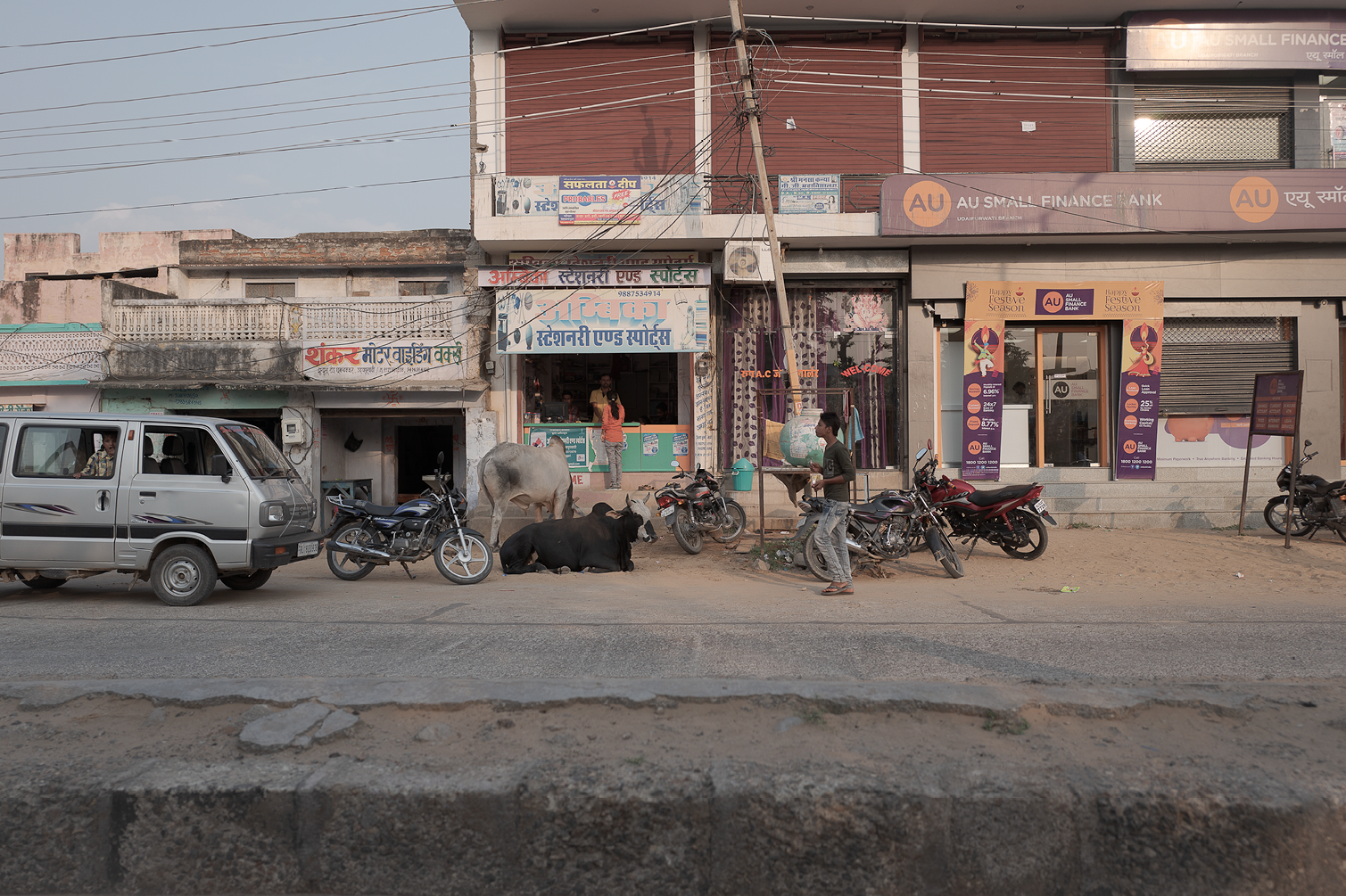 Une rue urbaine avec des bâtiments commerciaux, des motos, un véhicule et des vaches se reposant sur le trottoir. Plusieurs personnes sont présentes, dont une passant à côté d'une vache couchée, et une autre à la fenêtre du magasin.