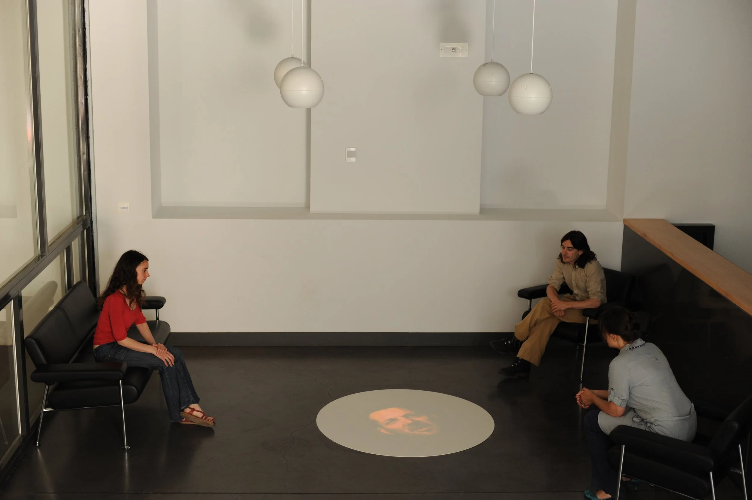 Salle d'attente avec cinq personnes assises sur des chaises noires en cercle. Il y a un plafond blanc avec trois lampes suspendues et un mur blanc avec des prises électriques.