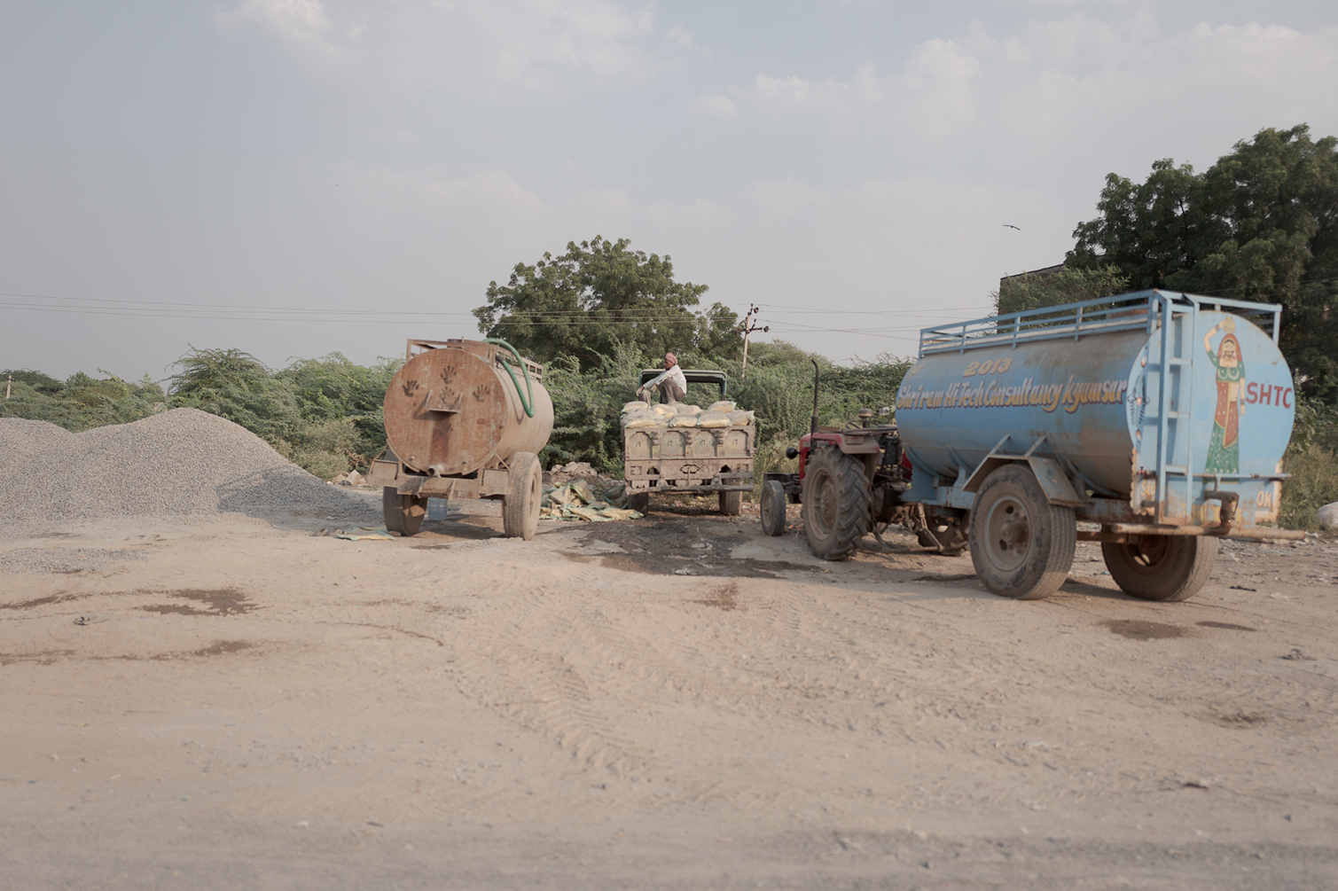 Travaux de construction ou de réparation, avec deux camions-citernes, un tracteur et un homme travaillant, sur une route en zone rurale, entourée d'arbres et de cabanons, sous un ciel partiellement nuageux.