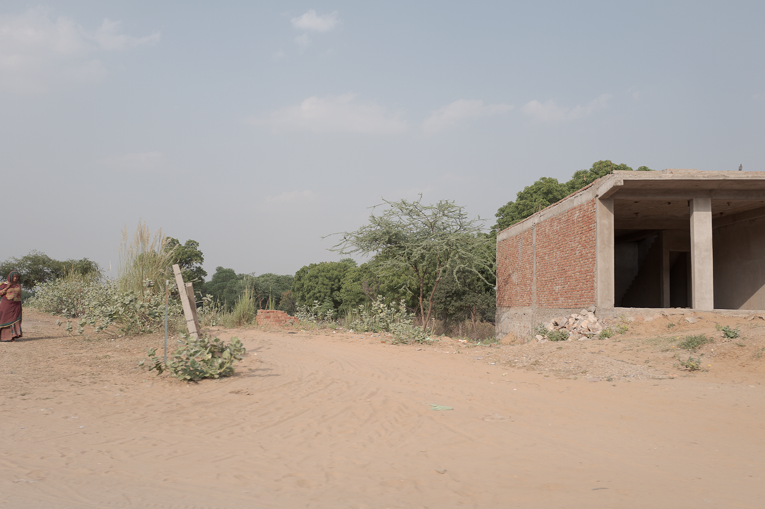 Une maison en construction en brique sans fenêtre, située en zone rurale avec quelques arbres et végétation sèche, et une femme portant un vêtement long à gauche.
