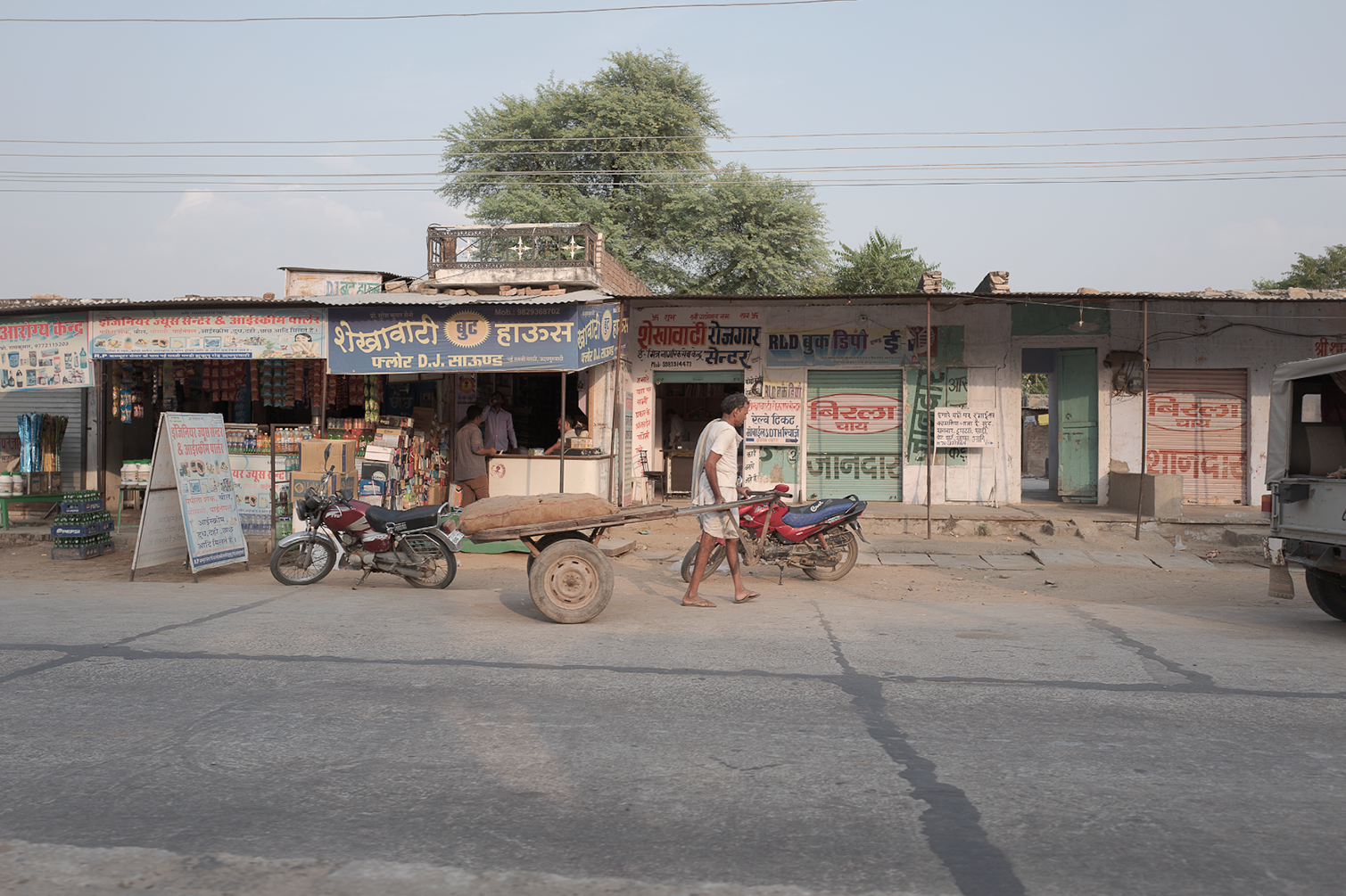 Une rue avec des boutiques en façade, un homme poussant une charrette avec du bois, deux motos garées devant, et une voiture à droite.