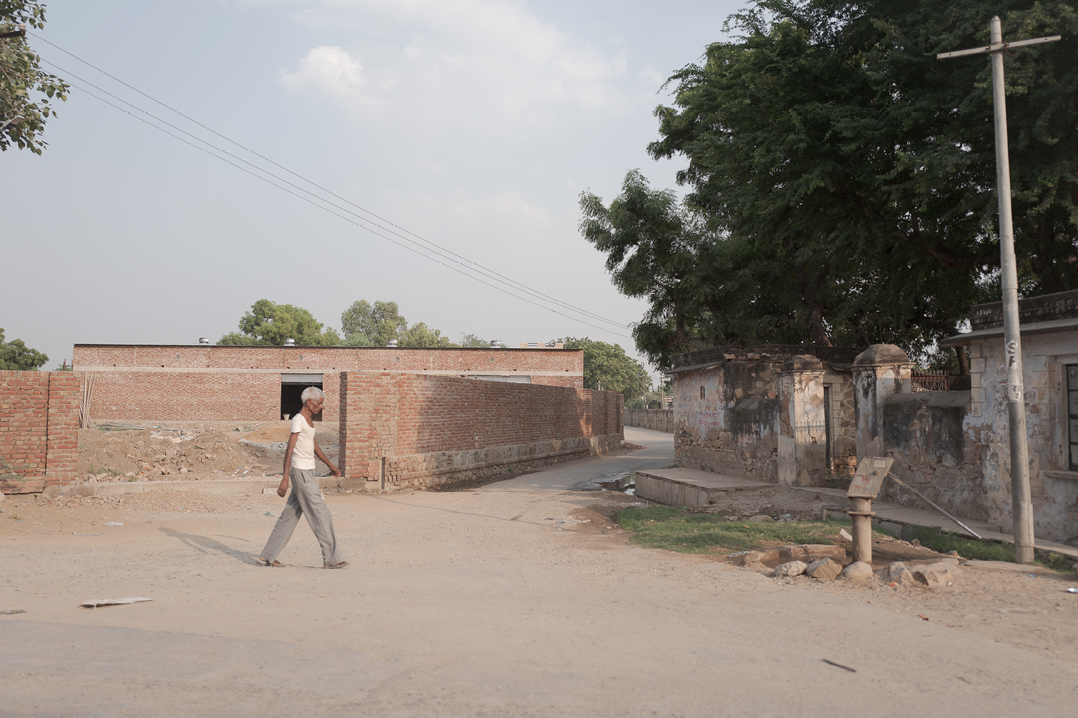 Une femme âgée avec des cheveux gris, portant un t-shirt blanc et un pantalon gris, marche sur une route de terre dans un quartier résidentiel avec des murs en briques et en pierre, des arbres verts et un panneau d'information à droite.
