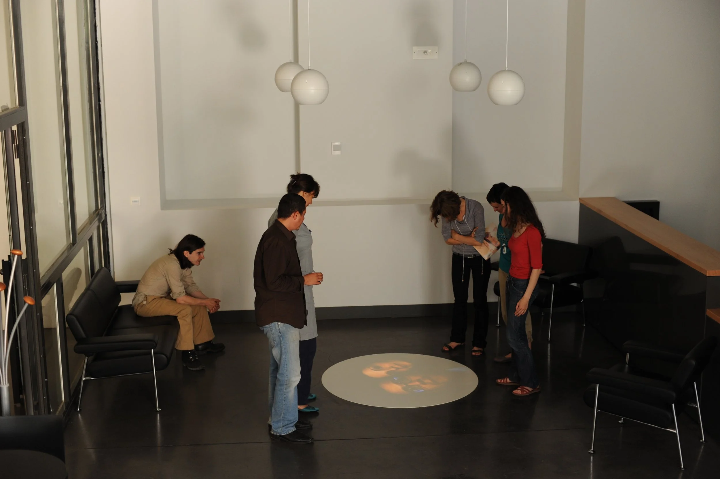 Groupe de personnes debout dans une salle moderne, regardant un objet ou une présentation sur une table, avec un canapé noir et des lampes suspendues au plafond.