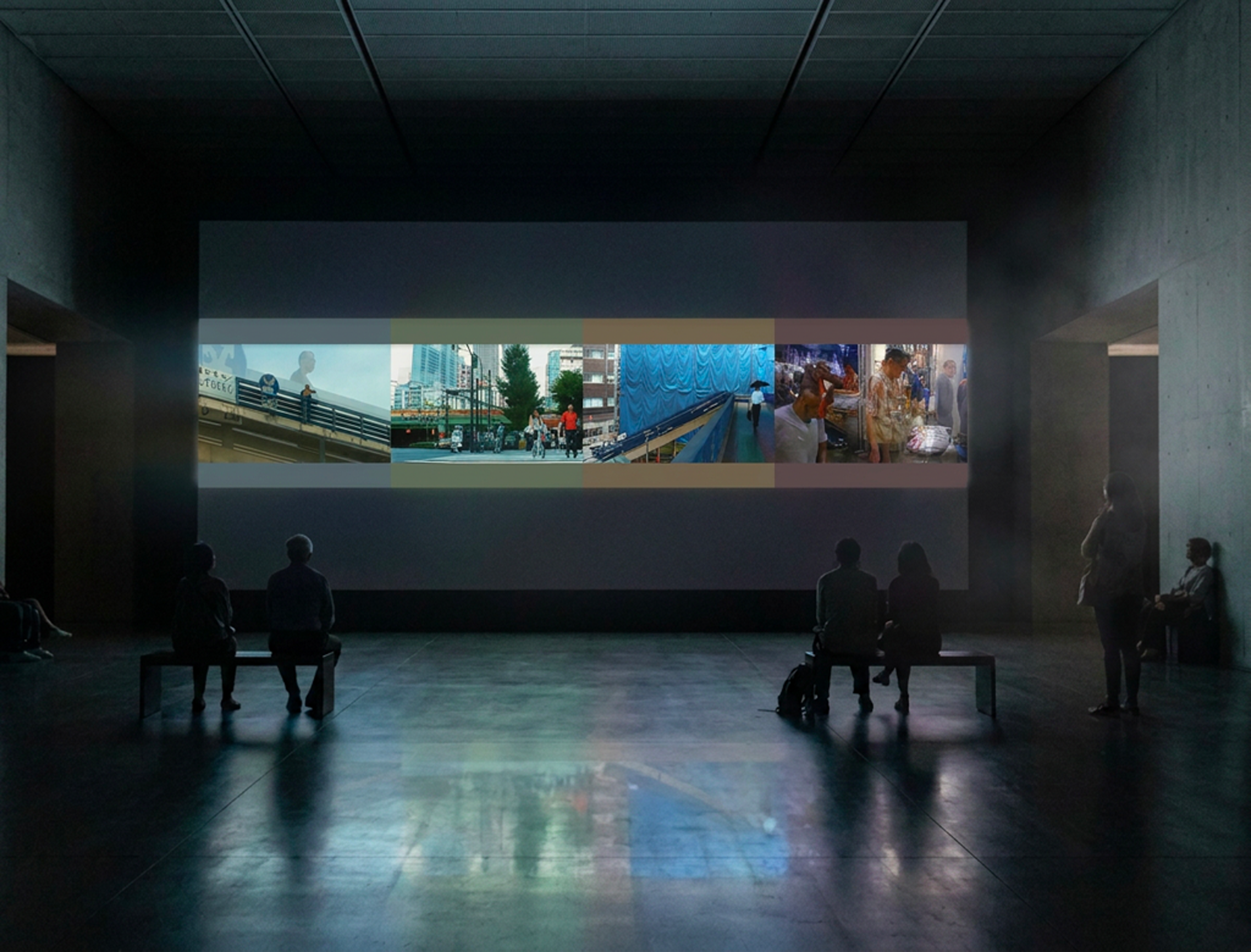 Salle sombre avec un grand écran affichant une vidéo de scènes urbaines et de personnes, des silhouettes de spectateurs assis.