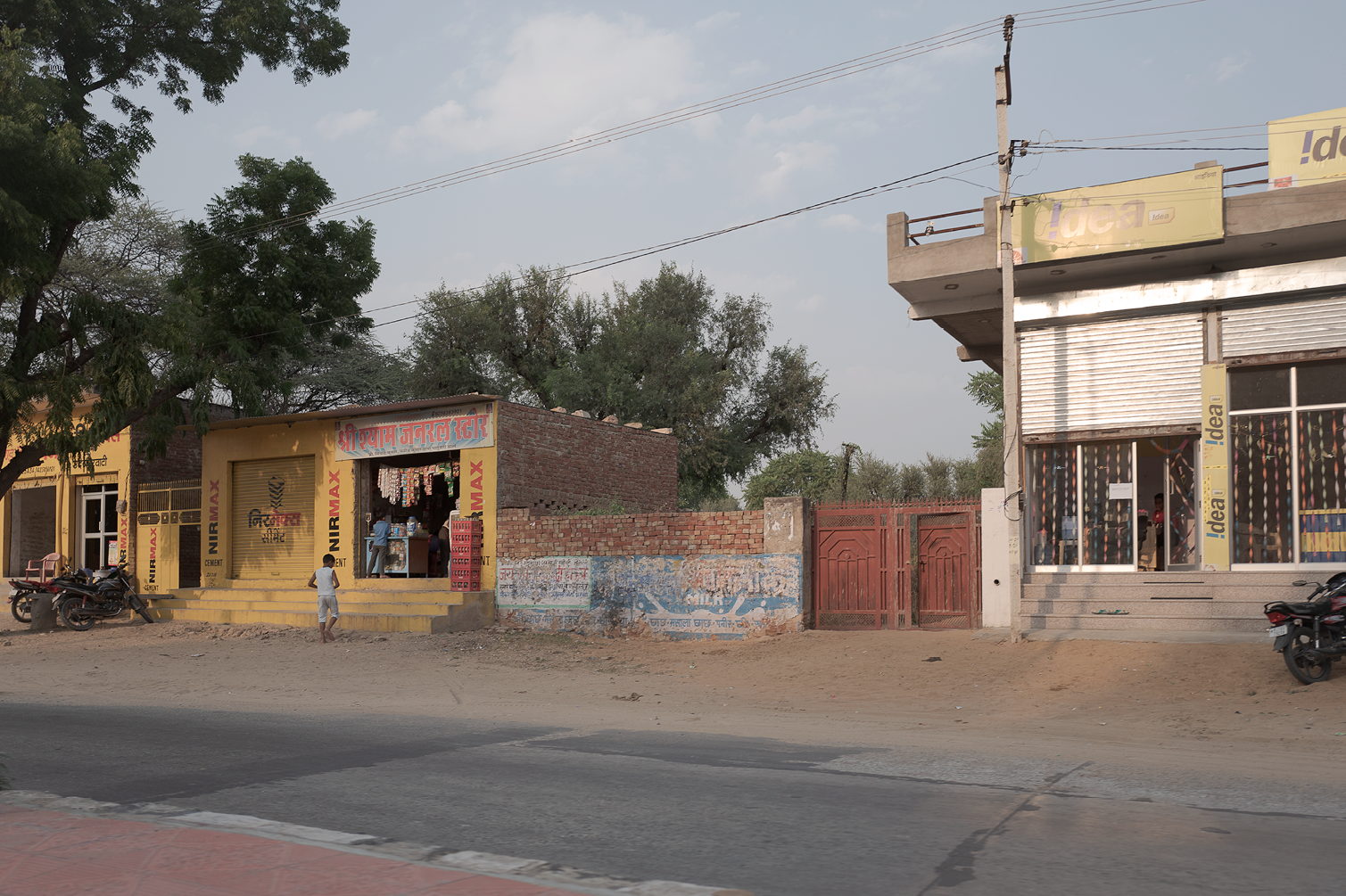 Rue dans une zone commerciale avec des boutiques, y compris un magasin avec la enseigne en hindi, et un autre avec la enseigne 'Idea'. Motos garées sur le côté de la route, et une barrière en métal rouge. Arbres et ciel avec quelques nuages en fond.