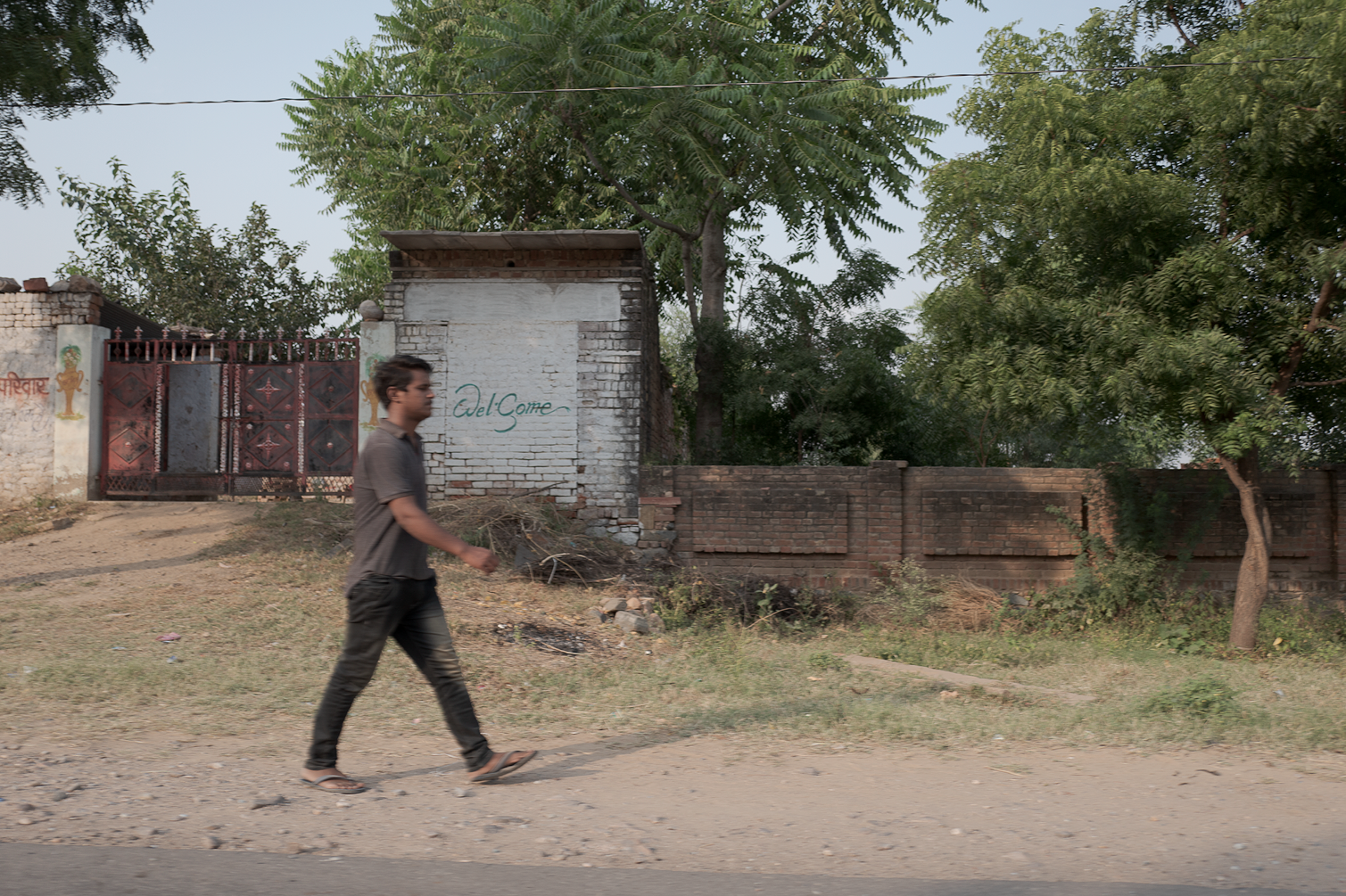 Un jeune homme marche le long d'une route en terrain poussiéreux avec une clôture en brique et un mur en pierre derrière lui, où il est écrit 'Del Come' en vert. Il y a des arbres verts et un portail en métal rouge à gauche.