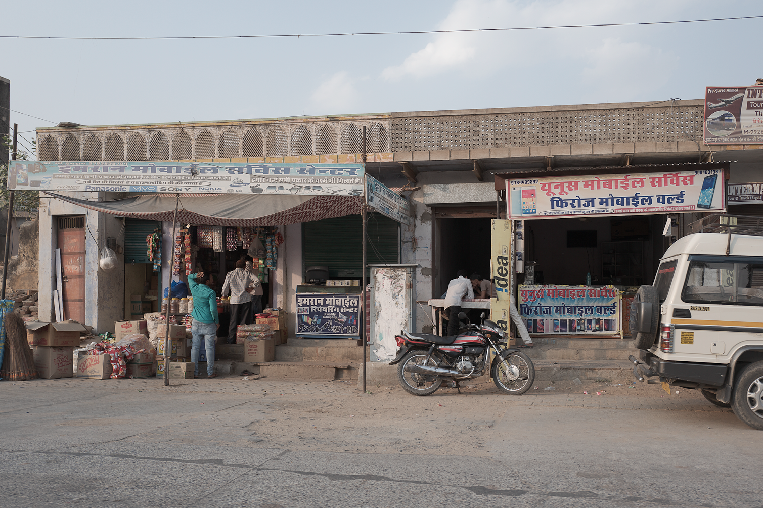 Vitrine d'un magasin de téléphones mobiles avec des affiches publicitaires en hindi, des clients achetant des produits, une moto garée devant le magasin, et un véhicule blanc stationné sur la route poussiéreuse.