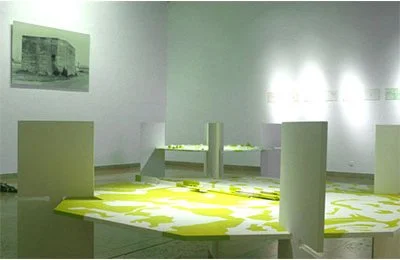 Exposition d'art contemporain dans une galerie, avec murs blancs, œuvres modernes, et une table en centre avec une peinture verte sur un mur.