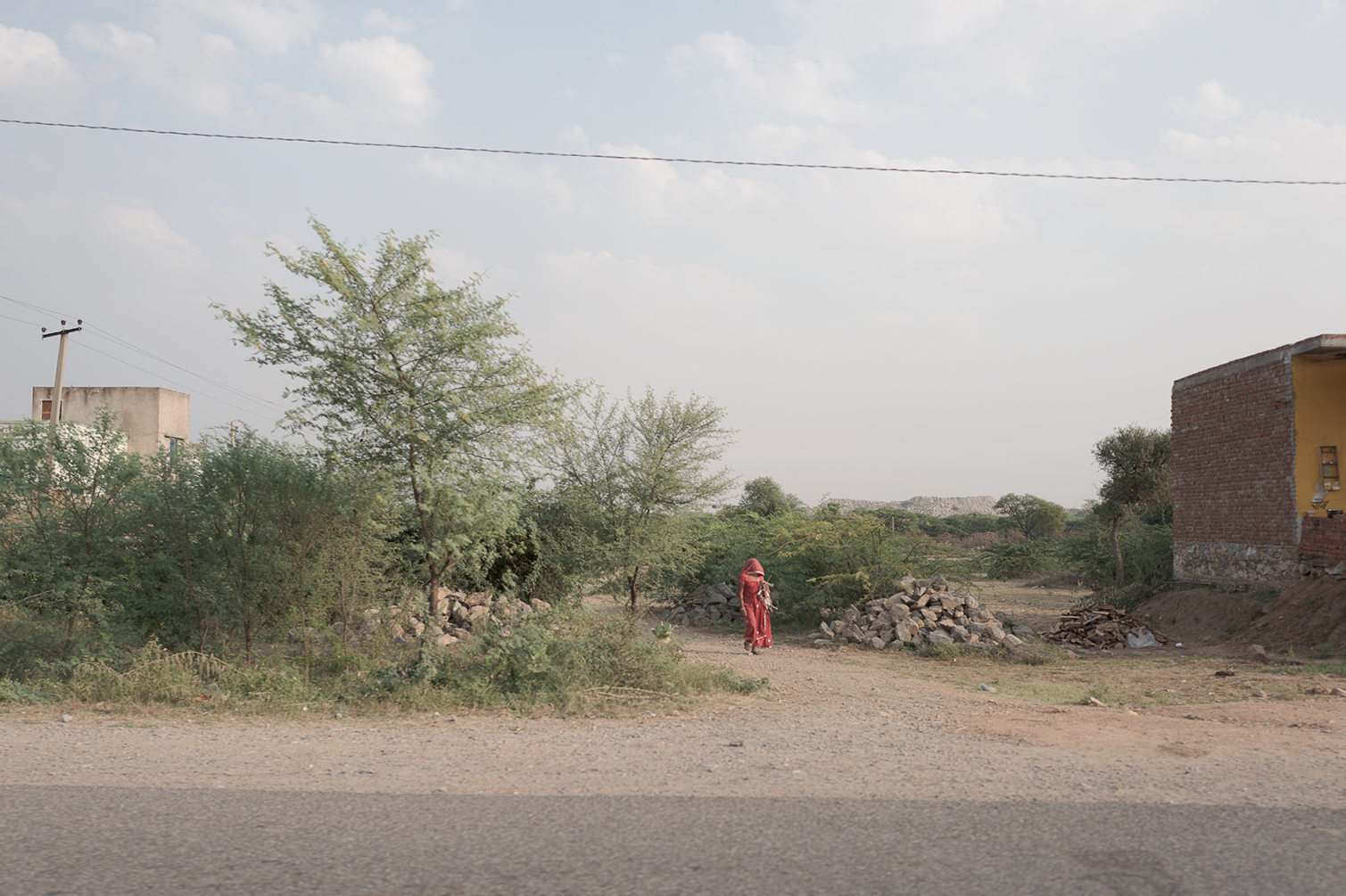 Une femme marchant le long d'une route dans une zone rurale peu développée, avec des arbres, des piles de pierres et des bâtiments en arrière-plan, sous un ciel partiellement nuageux.