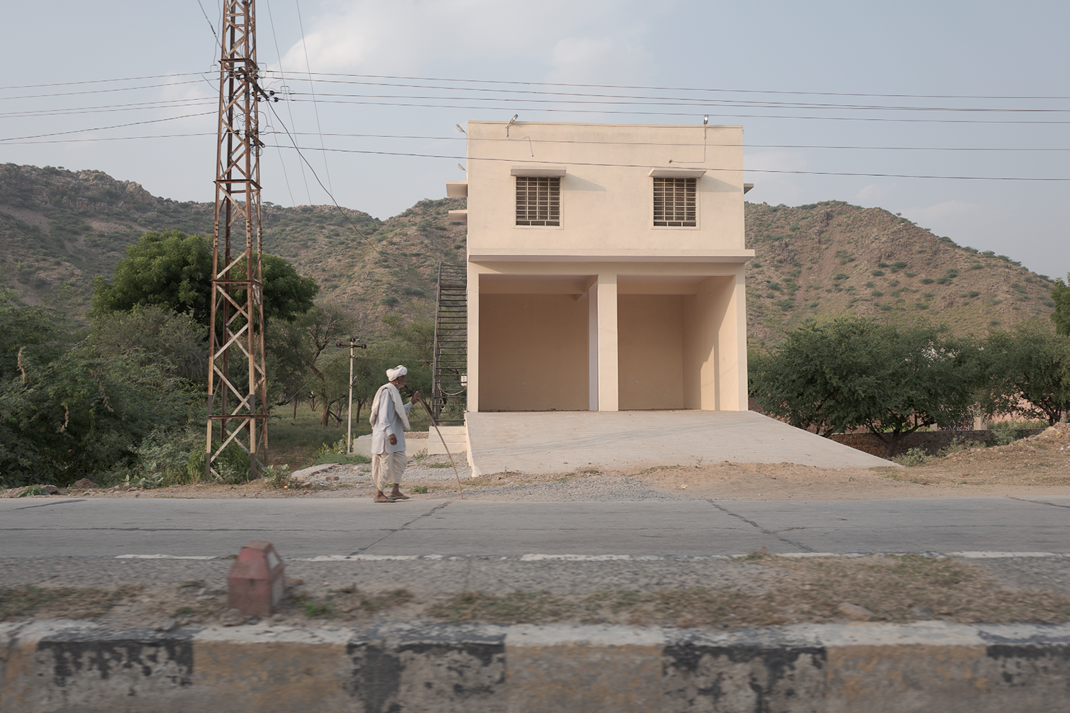 Une maison inachevée en construction, avec une montée en pente pour la voiture. Un homme âgé avec une turban blanc se tient sur le bord de la route, utilisant une canne, en face de la maison. Des montagnes en arrière-plan et des arbres autour.
