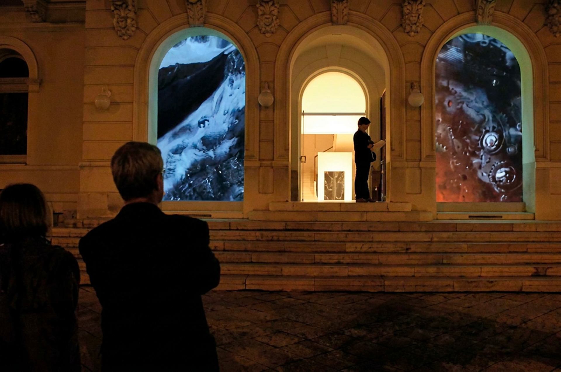 Une galerie d'art en plein air avec des visiteurs regardant des images projetées sur des fenêtres en pierre, dont l'une montre une cascade, et une autre des engrenages mécaniques.