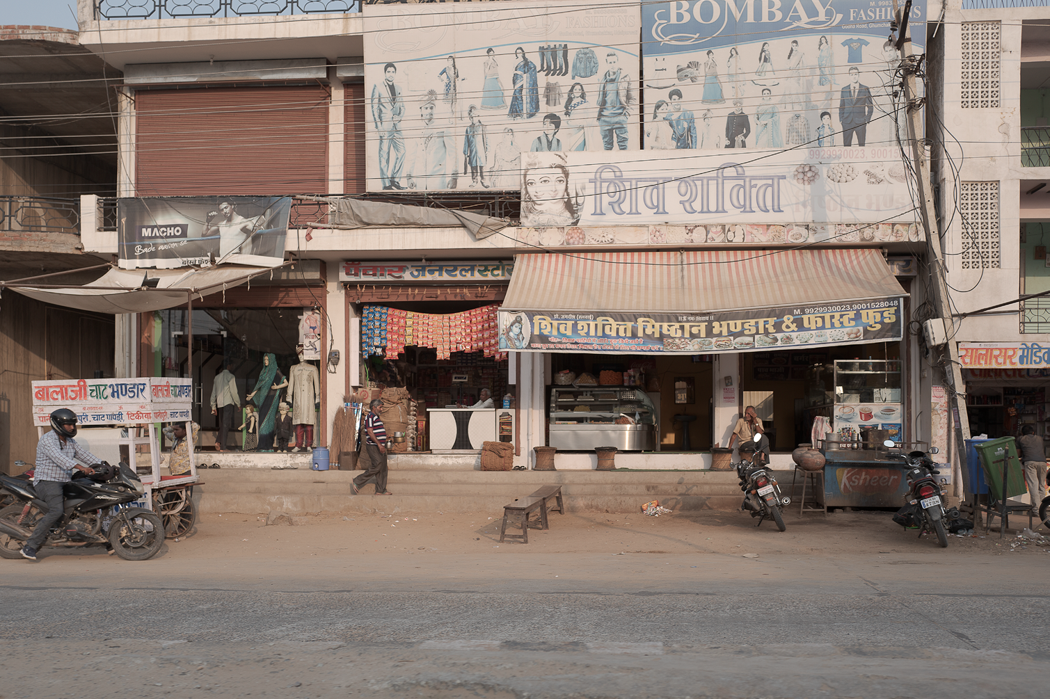 Un aperçu de la façade d'un marché ou d'un magasin en Inde avec des enseignes en hindi, plusieurs motos garées devant, des personnes marchant et un mannequin exhibant des vêtements. La scène est prise depuis la route, en milieu urbain.