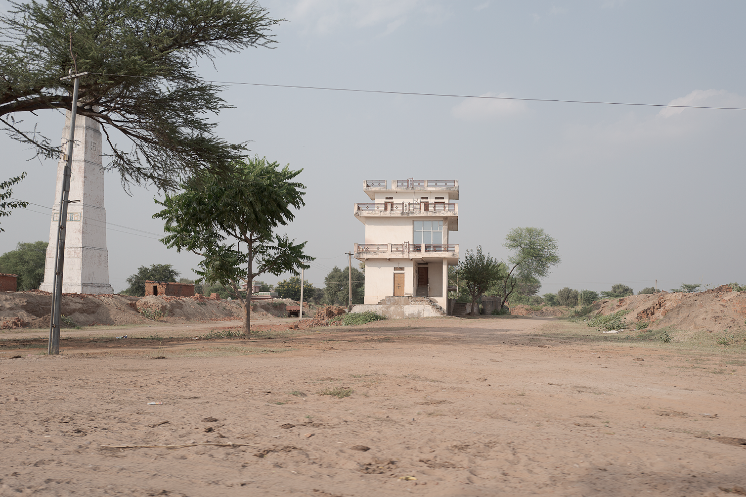 Une maison blanche à trois étages avec un balcon à chaque niveau, située dans un paysage rural sec avec quelques arbres, et un pylône électrique à gauche.