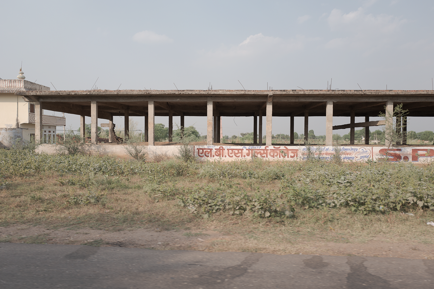 Bâtiment abandonné avec structures en béton, fenêtres ouvertes, graffitis en hindi sur le mur en bas, végétation sauvage devant, ciel clair avec quelques nuages.