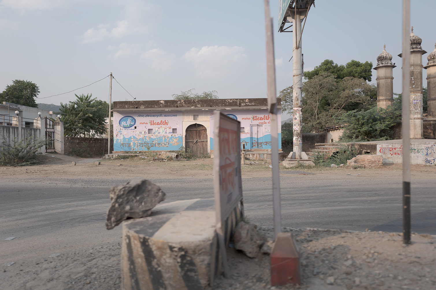 Route poussiéreuse avec un panneau de signalisation, une vieille maison avec un mur peint présentant des publicités et des textes en hindi, et quelques arbres. Le ciel est clair avec quelques nuages.