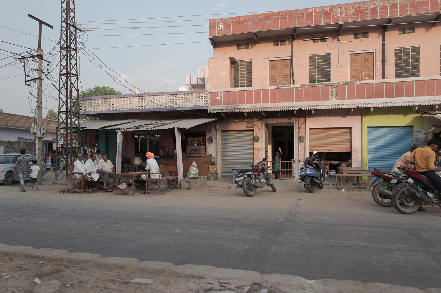 Une rue urbaine avec un bâtiment rose à plusieurs étages, des scooters garés devant, et des personnes assises sur des bancs en train de parler ou attendre. Quelques magasins avec des volets roulants, et des passants.