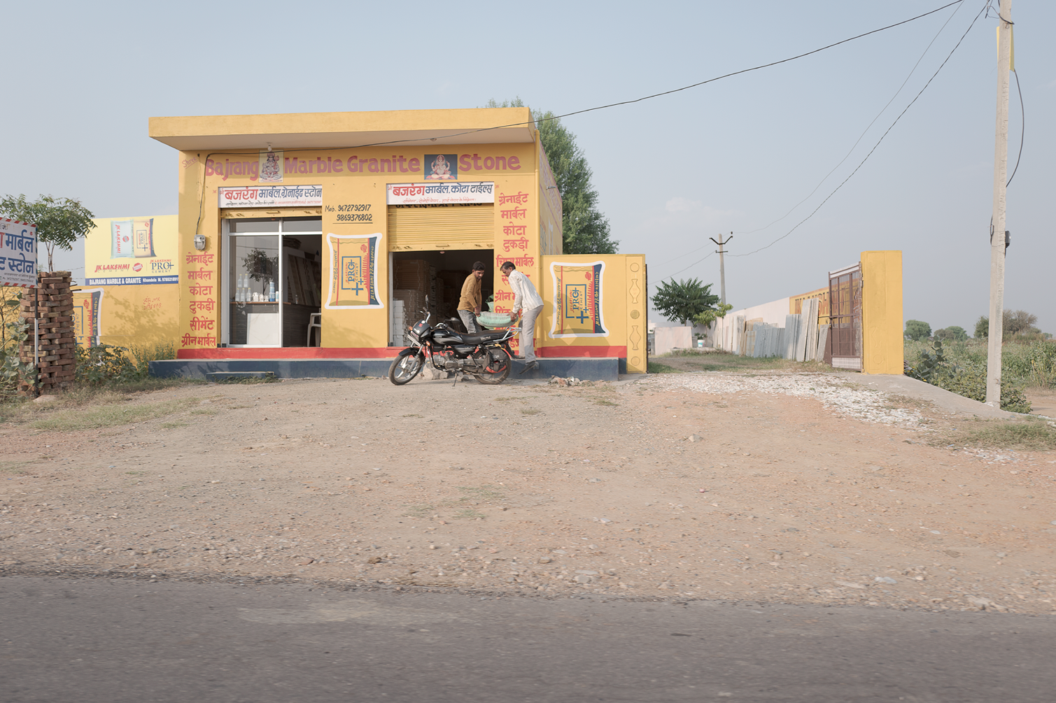 Une petite boutique de pierre, marbre et granit en Inde avec deux hommes discutant devant l'entrée, une moto garée devant, derrière une porte en métal et un panneau avec des annonces, le tout sous un ciel clair.