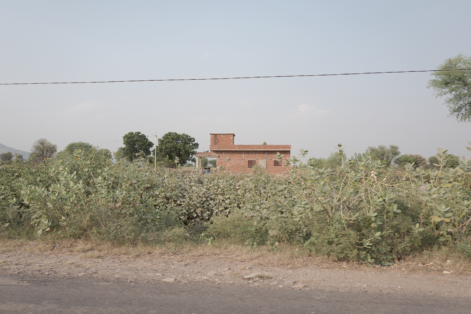 Une maison en briques rouges dans un paysage rural avec des buissons et des arbres, sous un ciel clair.