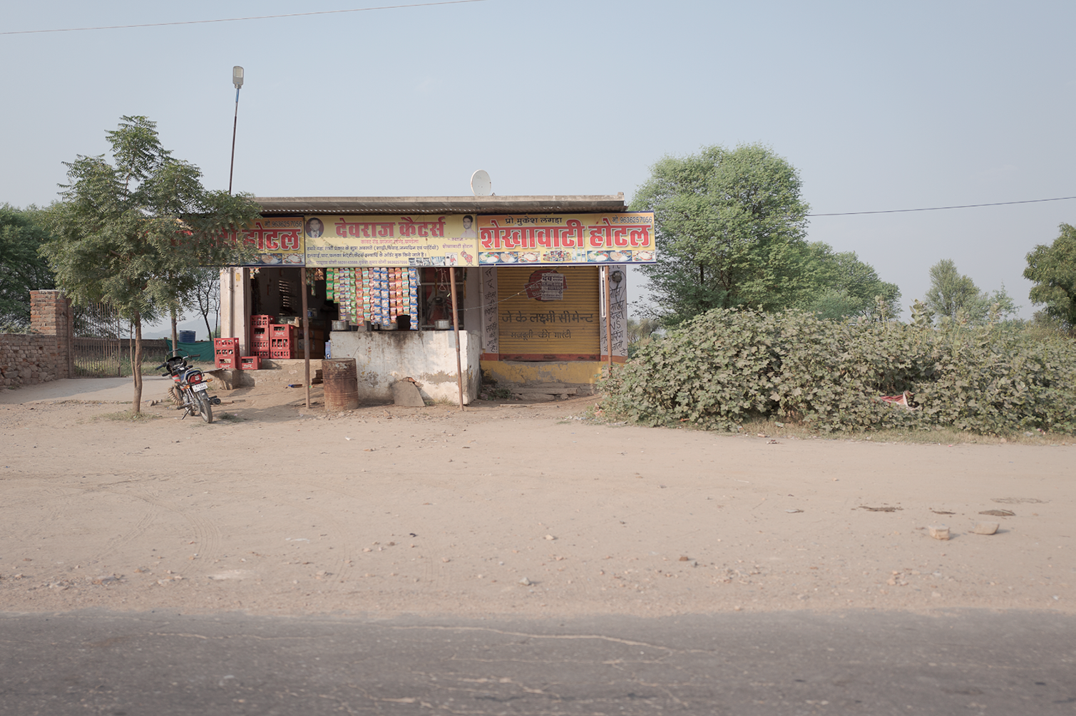 Un petit magasin ou stand avec des panneaux d'affichage en hindi, situé dans un environnement sec avec peu de végétation, et une moto garée devant. La façade du magasin comporte des articles suspendus et des caisses en plastique.
