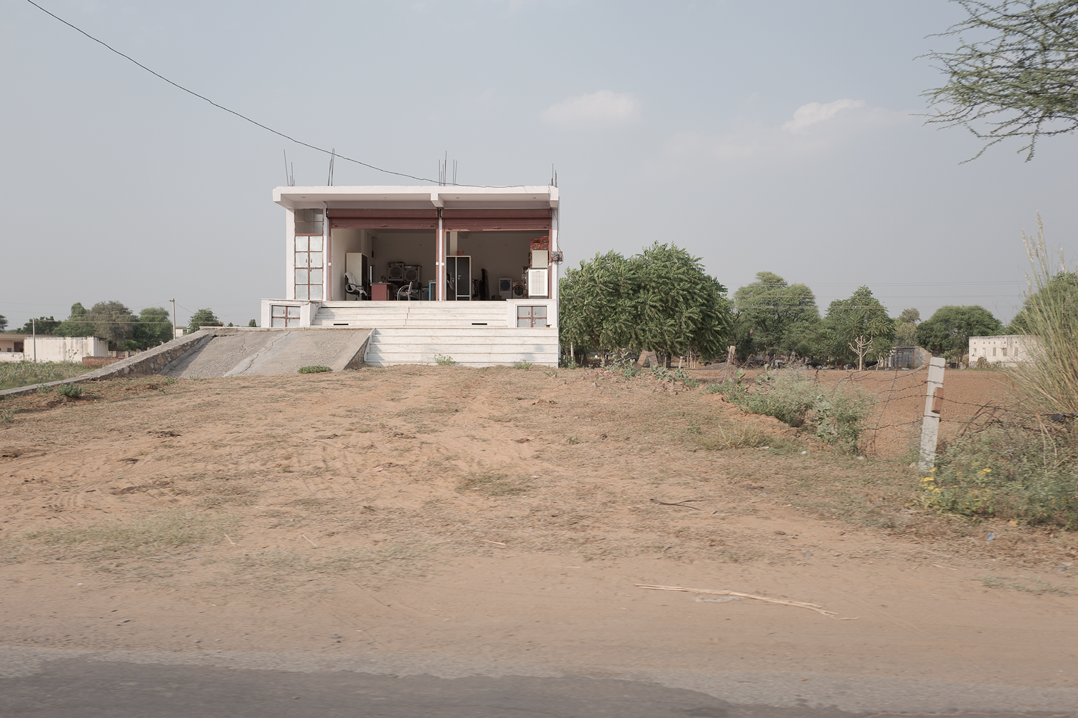 Une maison moderne blanche en hauteur avec une demi-verrière et un escalier blanc, située dans un environnement rural avec du terrain sec et peu de végétation.