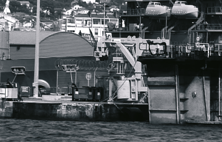 Vue d'un quai de port avec un navire militaire en arrière-plan, en noir et blanc.