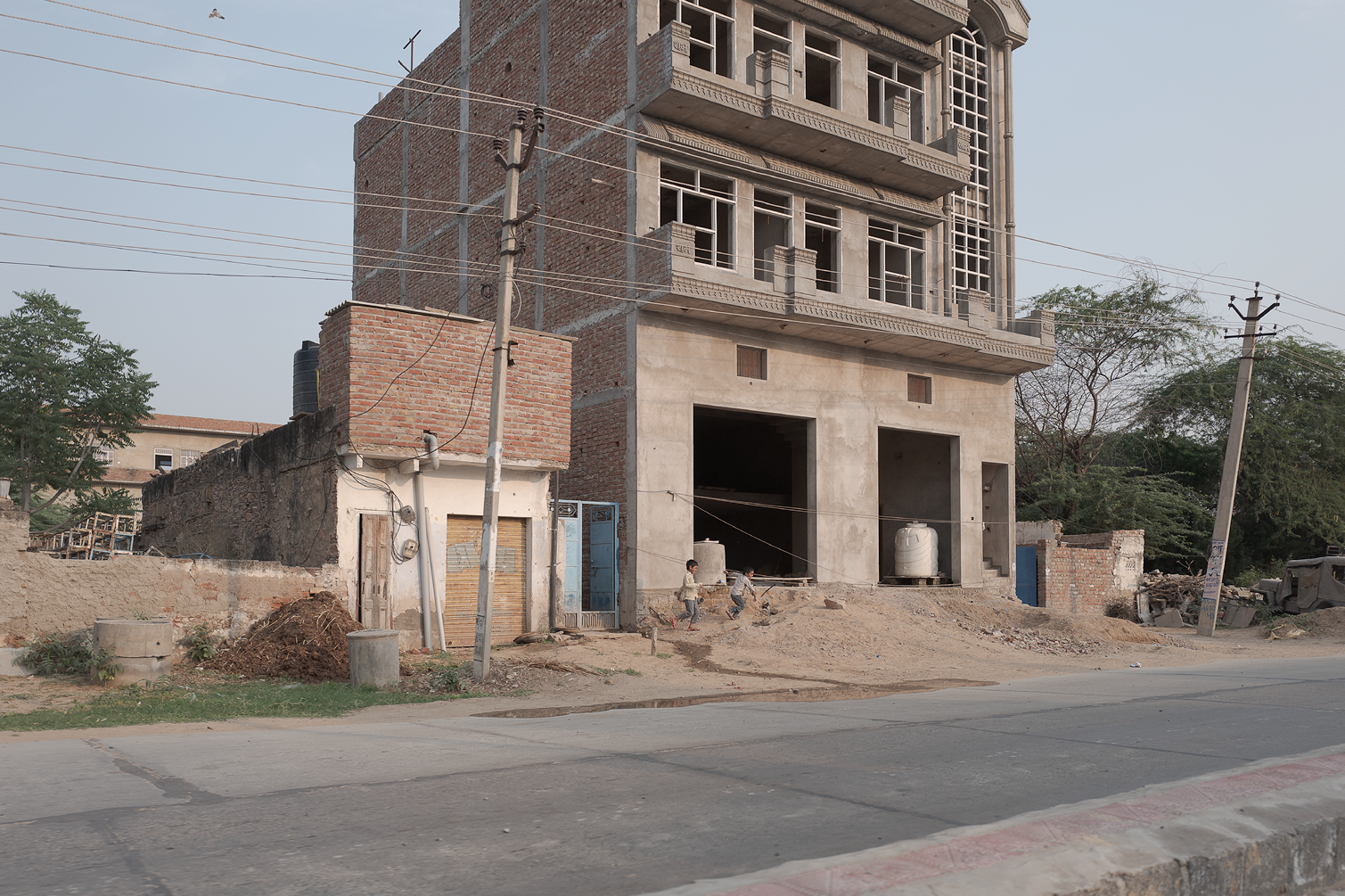 Une construction en cours avec un bâtiment à plusieurs étages sous construction, des enfants jouent devant un garage à porte en métal et un mur en brique, des poteaux électriques traversent la scène, et il y a des arbres au fond.
