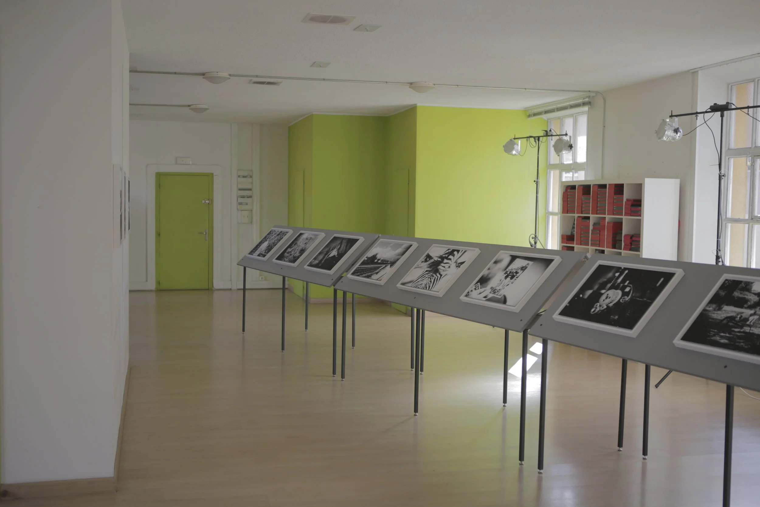 Une galerie d'art avec des photographies en noir et blanc exposées sur des tables, mur vert vif à l'arrière, fenêtres laissant entrer la lumière naturelle, et projecteurs suspendus au plafond.