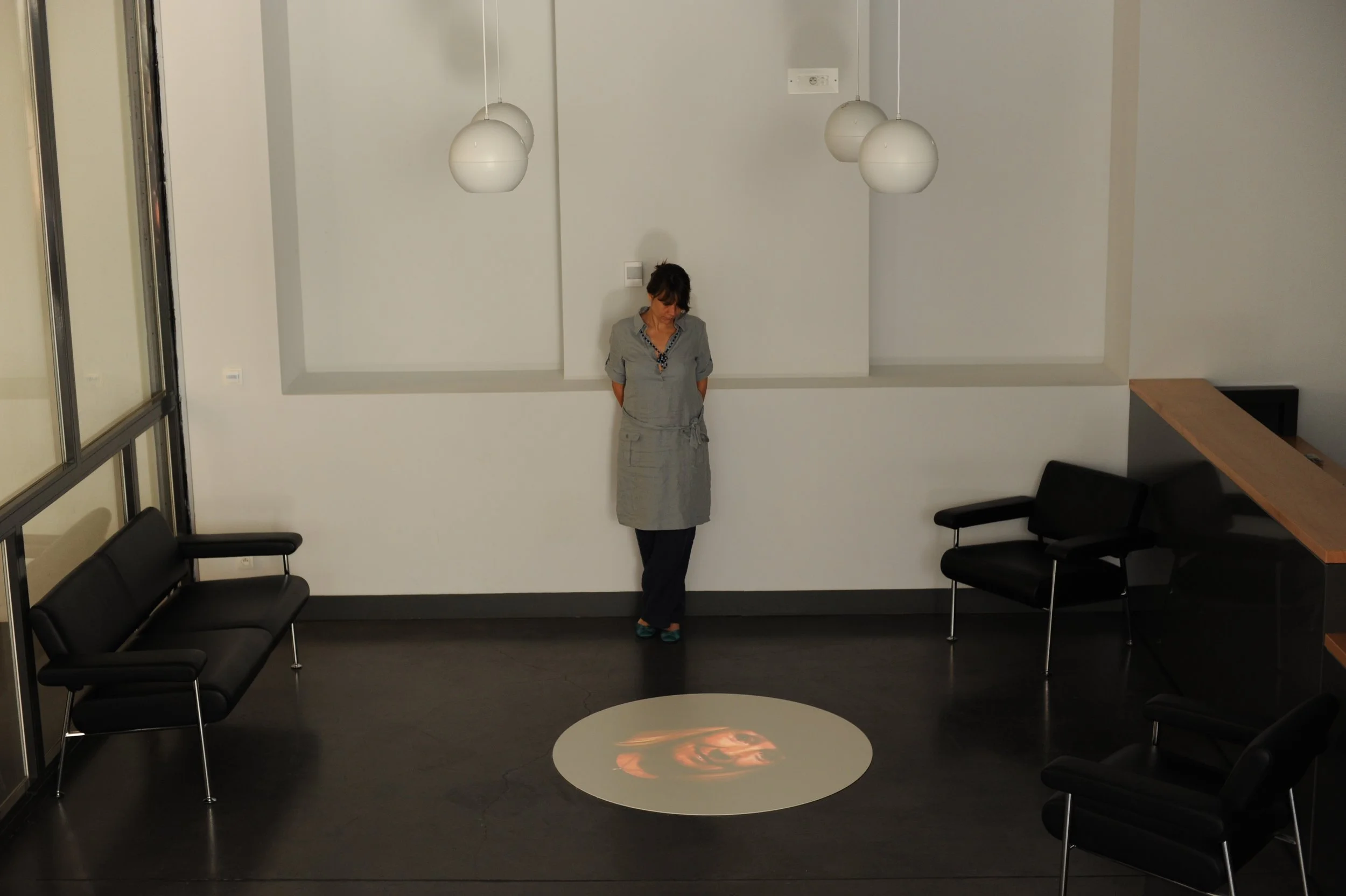 Une femme debout dans une salle d'attente, avec des chaises noires, un mur blanc, et un éclairage suspendu, un reflet de visage projeté au sol.