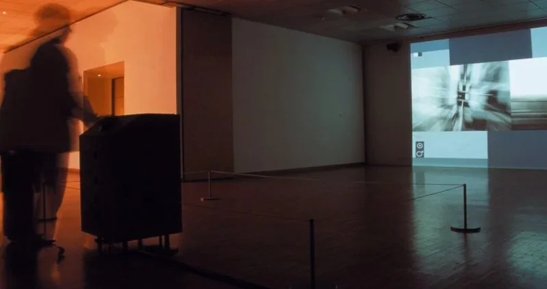 Une salle sombre avec une projection vidéo sur un mur blanc, et deux personnes floues qui regardent l'écran.