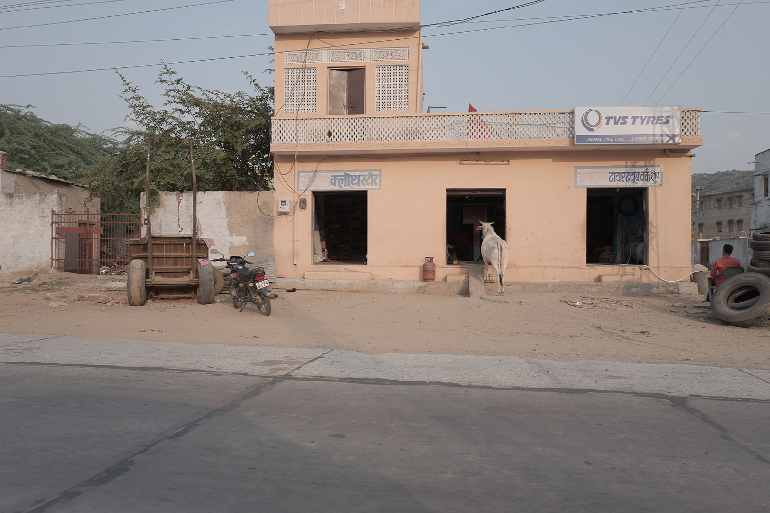 Une façade de bâtiment beige avec deux fenêtres au rez-de-chaussée, une porte au centre, un espace de vente de pneus TVS Tyres, un cow-boy de dos, une moto et une charrette en bois à gauche, des pneus empilés à droite, un homme assis sur les pneus et