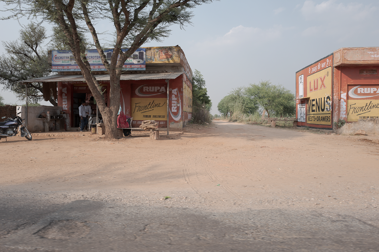 Une route en terre dans un village avec deux boutiques en pierre et panneaux publicitaires, quelques personnes et une moto sont visibles.