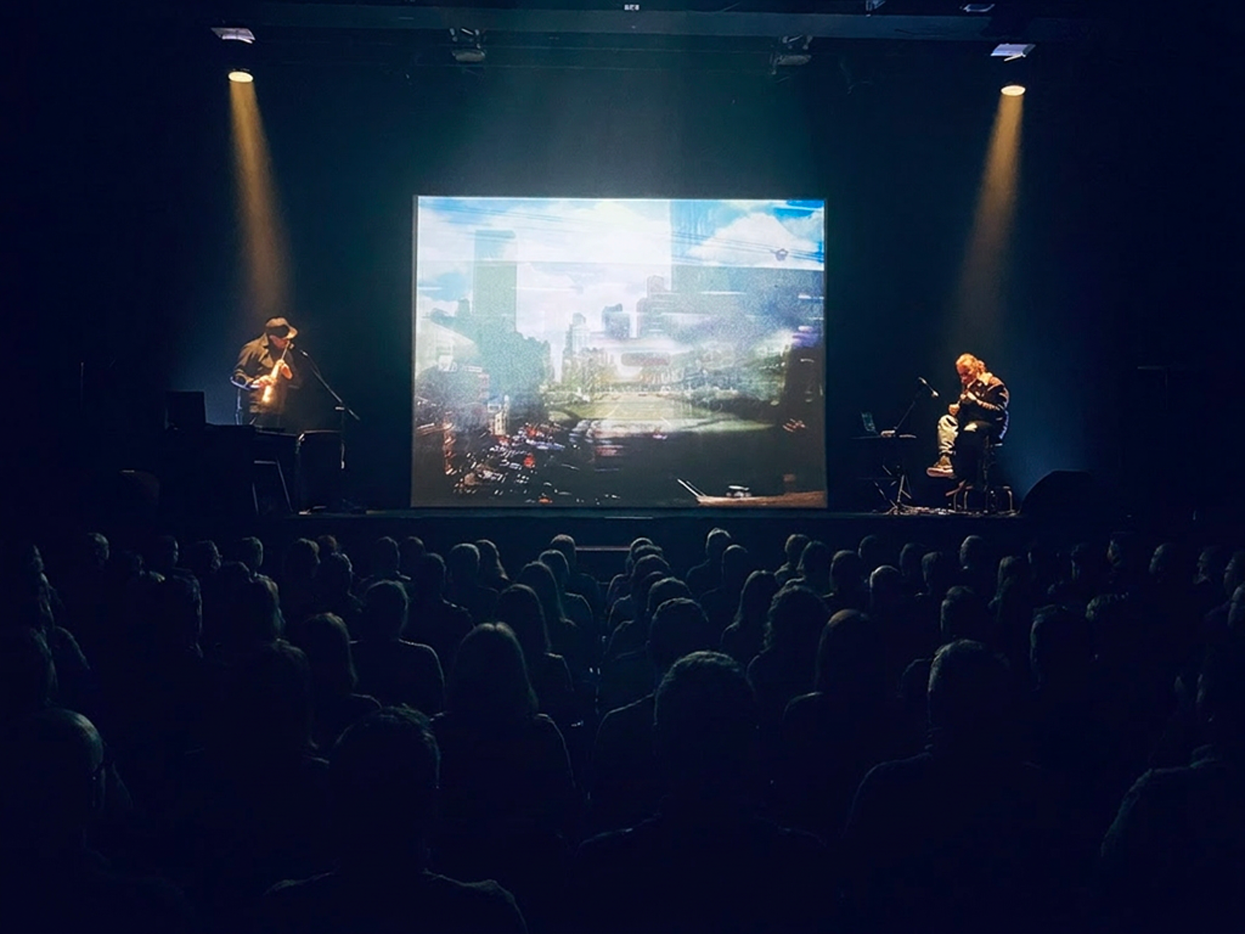 Une scène de spectacle avec deux musiciens, un à gauche jouant d'un instrument à vent et à droite jouant de la guitare, sur scène devant un grand écran affichant une image urbaine avec des bâtiments et la circulation.