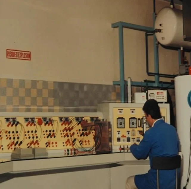 Un homme en blouse bleue travaillant avec des équipements électriques dans un laboratoire ou une salle technique.