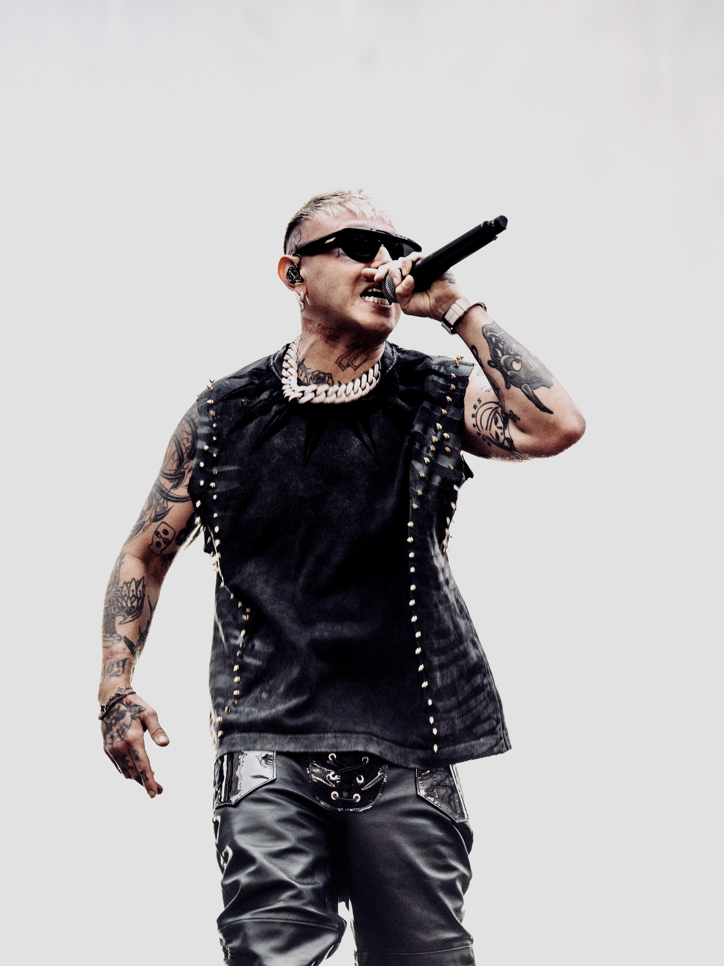 Un cantante con capelli corti e tatuaggi, indossa occhiali scuri e una collana di perline bianche, canta con un microfono in mano a un concerto.