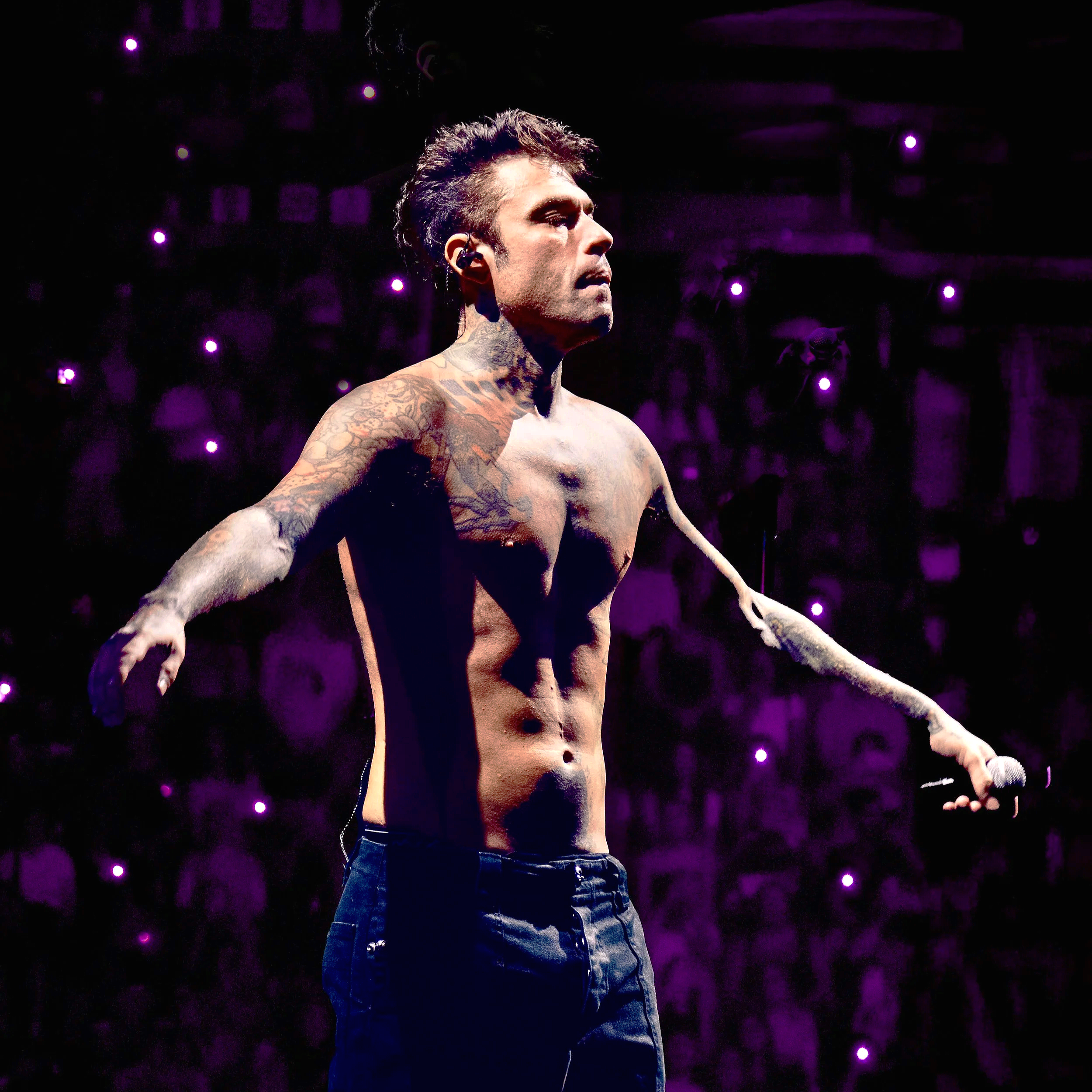 Un uomo con tatuaggi al petto e alle braccia indossa orecchini, senza camicia e con i jeans, in uno sfondo scuro con luci viola, sembra essere in un ambiente di spettacolo o concerto.