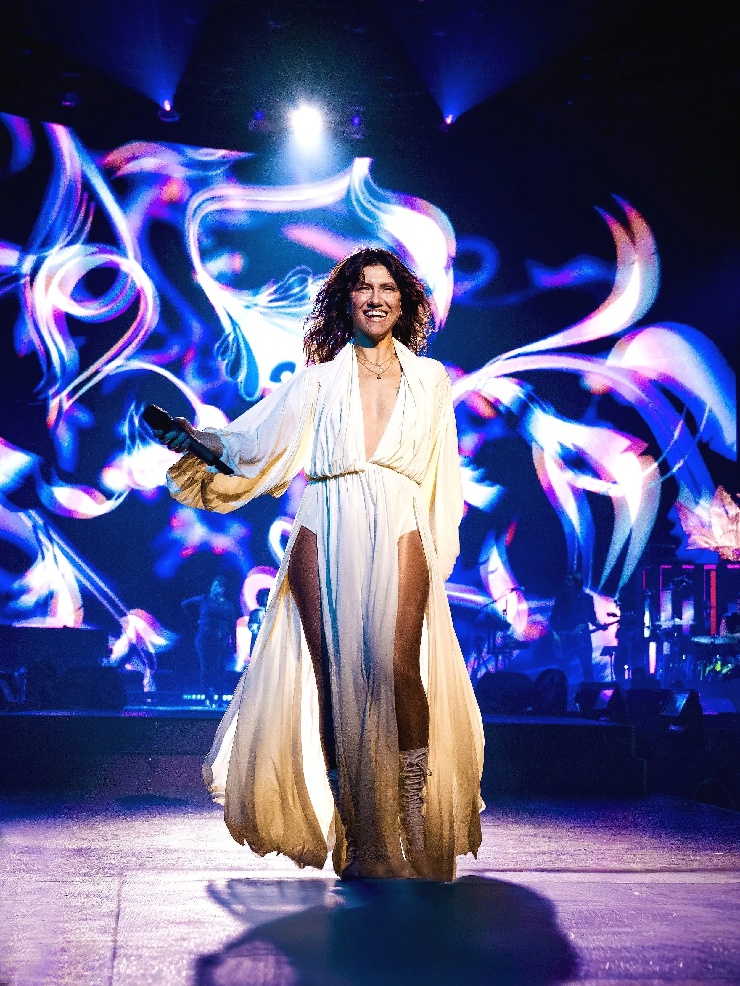 Una donna con un abito lungo e spacco alto e ciabatte con tacco si trova al centro di un palco, con uno sfondo di luci e immagini in movimento che creano un effetto artistico, mentre sorride e tiene un microfono in mano.